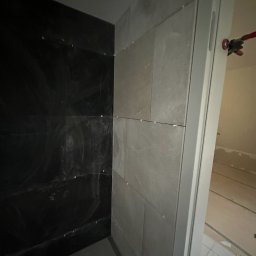 Hydro-Fliz - Ściana łazienki w trakcie wykańczania: część z ciemnymi płytkami, część z jasnymi, widoczne krzyżyki dystansowe i narzędzie glazurnicze przy futrynie.