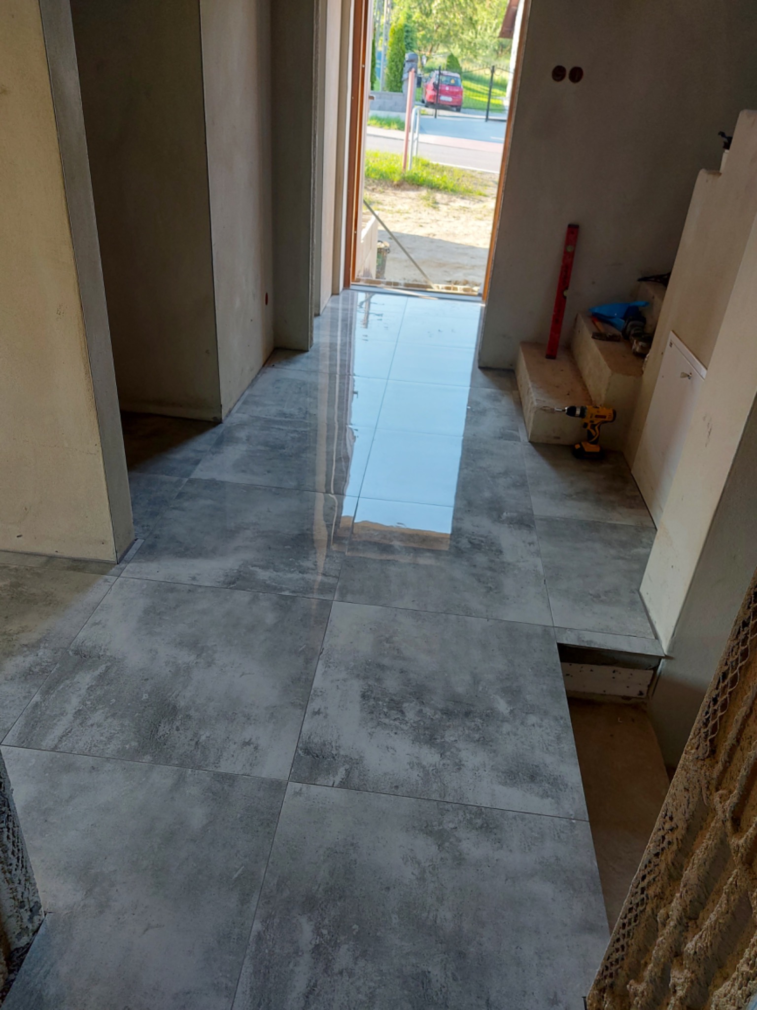 Ułożone, szare, matowe płytki podłogowe w korytarzu domu jednorodzinnego. Widoczny poziomica, wiertarka i częściowo ułożone schody.
