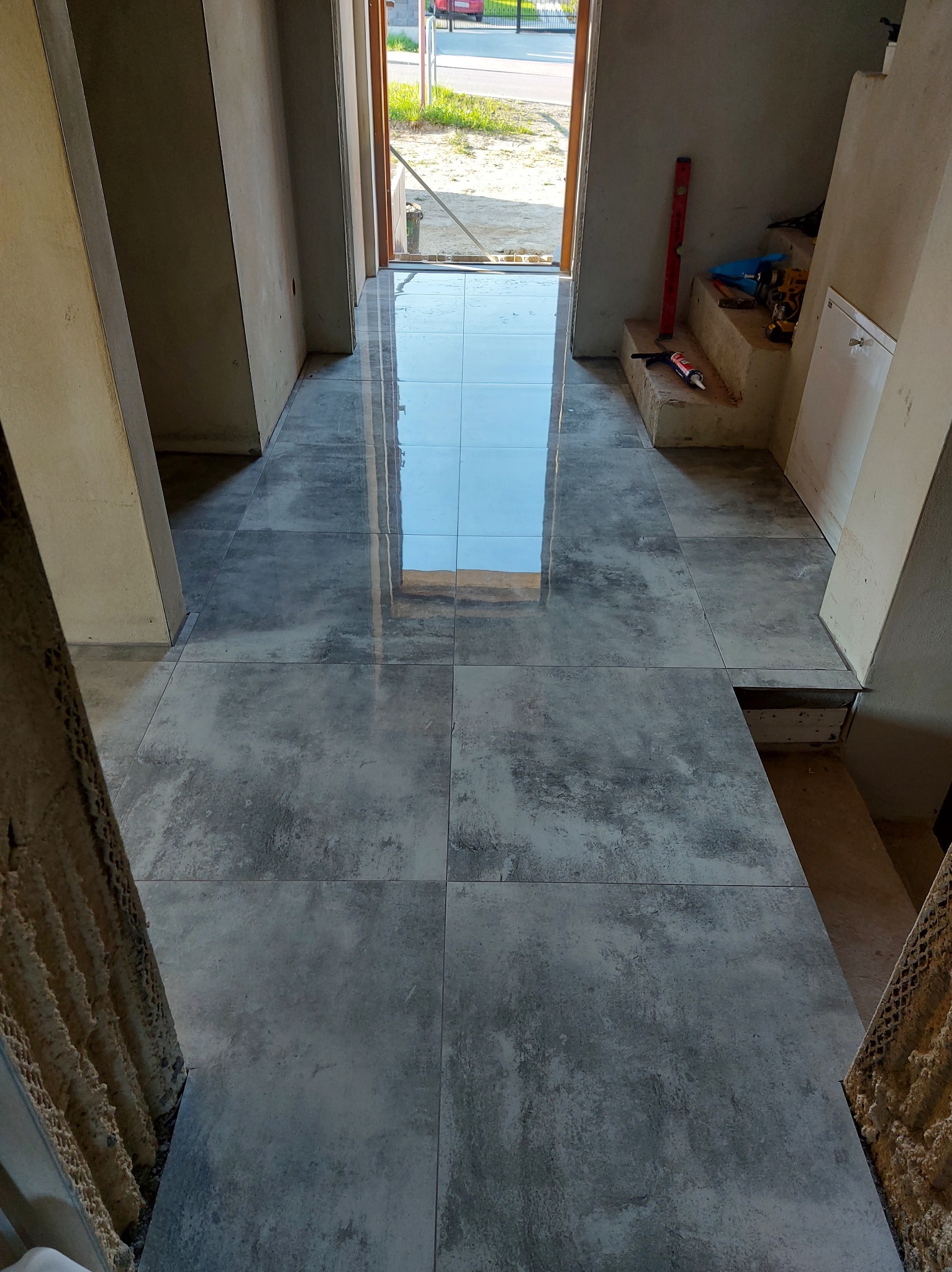 Ułożona podłoga z szarych, matowych płytek w korytarzu, z odbiciem nieba w połysku. Widoczne narzędzia pracy i poziomica. W tle otwarte drzwi wejściowe.