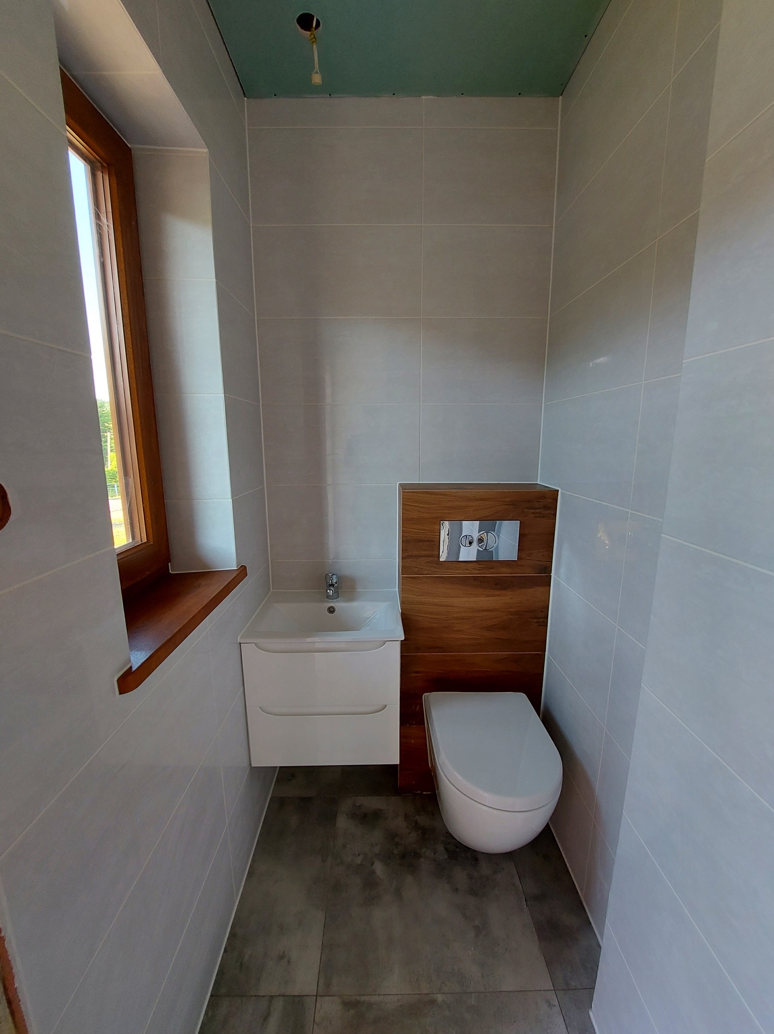 Jasna łazienka z drewnianymi akcentami: umywalka z szafką, podwieszana toaleta i okno z drewnianym parapetem. Szare płytki na ścianach i podłodze.