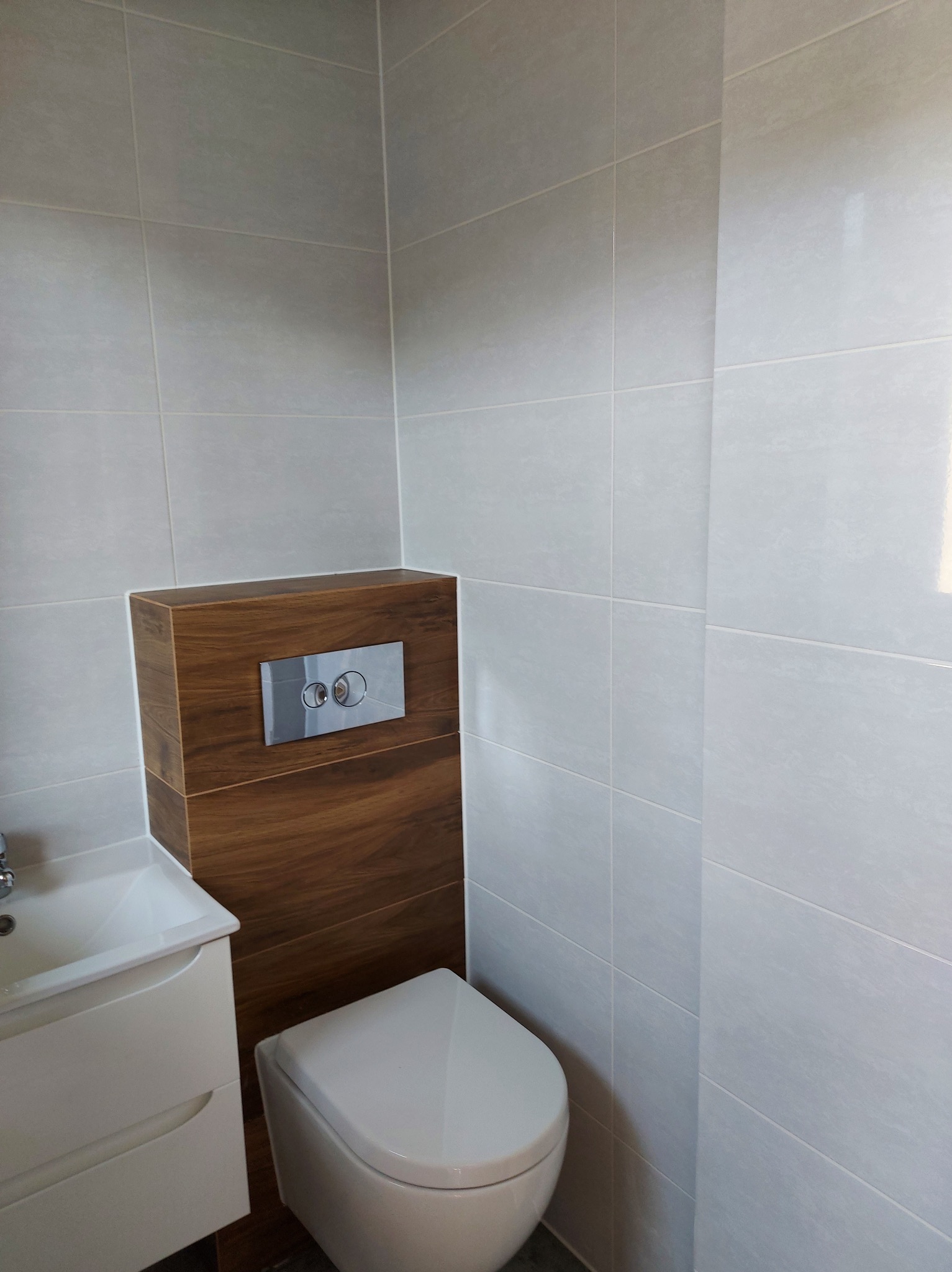 Nowoczesna łazienka z podwieszaną toaletą i umywalką, wykończona jasnymi płytkami i drewnopodobną zabudową stelaża WC. Minimalistyczny design.