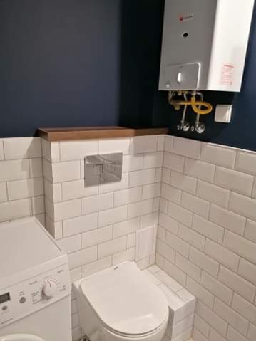 Wykończona łazienka z białymi, prostokątnymi płytkami, wiszącą toaletą i pralką. Na ścianie zamontowany piecyk gazowy i drewniana półka. Ciemna ściana w tle.