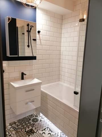 Biała łazienka z wanną obudowaną płytkami, umywalką z szafką i lustrem. Na podłodze wzorzyste płytki. Oświetlenie sufitowe i pod wanną.