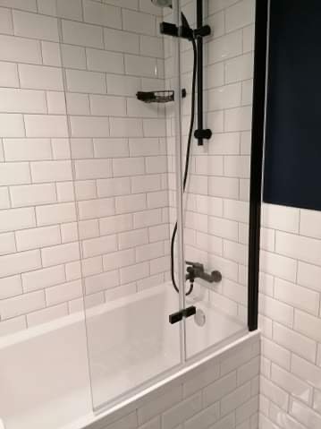 Biała wanna z czarną armaturą i szklanym parawanem nawannowym, obłożona białymi, prostokątnymi płytkami imitującymi cegłę. Nowoczesna łazienka.