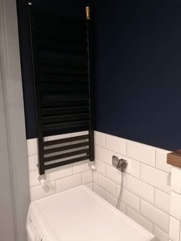 Nowoczesna łazienka z czarnym grzejnikiem drabinkowym na tle białych płytek i granatowej ściany. Widoczny fragment pralki. Minimalistyczny design.
