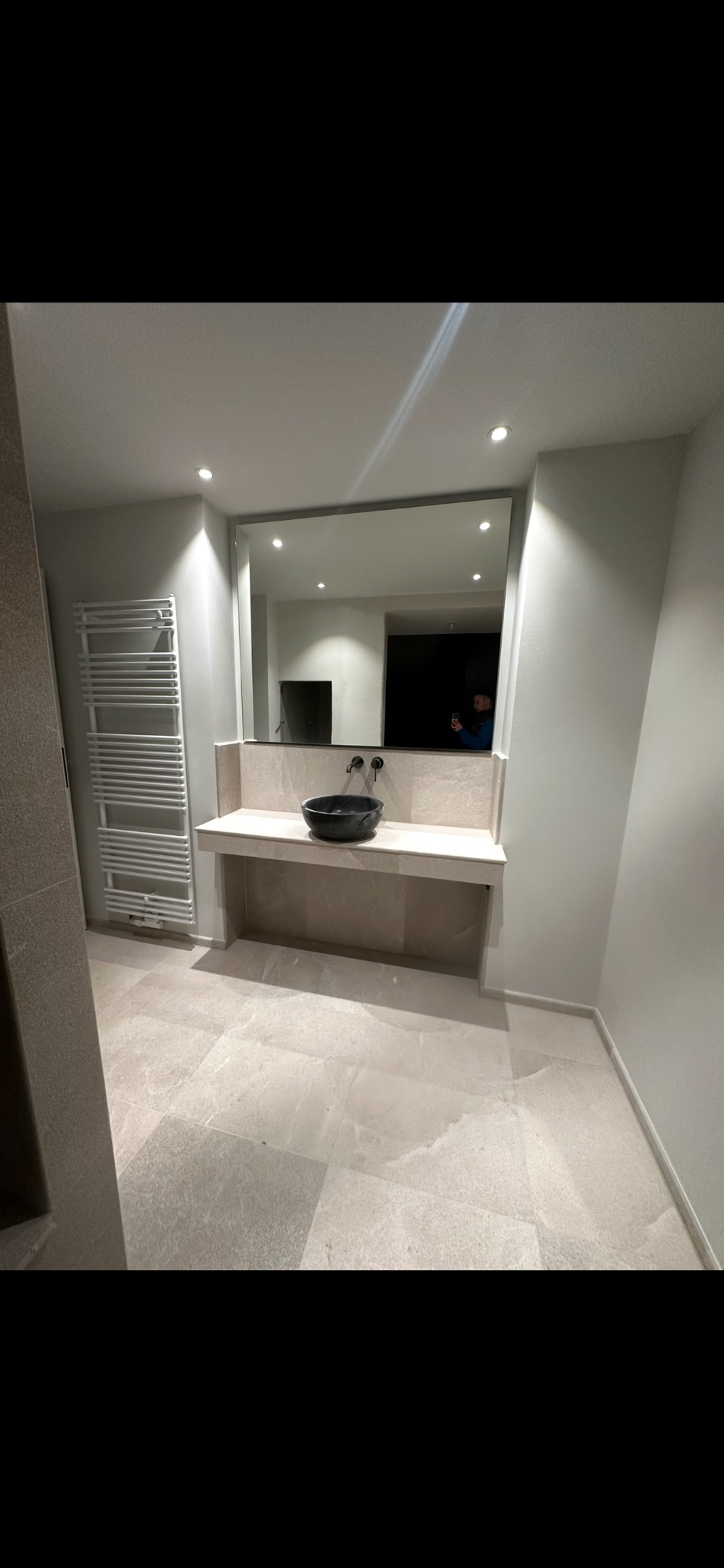 Wykończona łazienka w jasnych odcieniach beżu z czarną umywalką na blacie. Na ścianie grzejnik drabinkowy i duże lustro. Widoczne płytki podłogowe i oświetlenie punktowe.