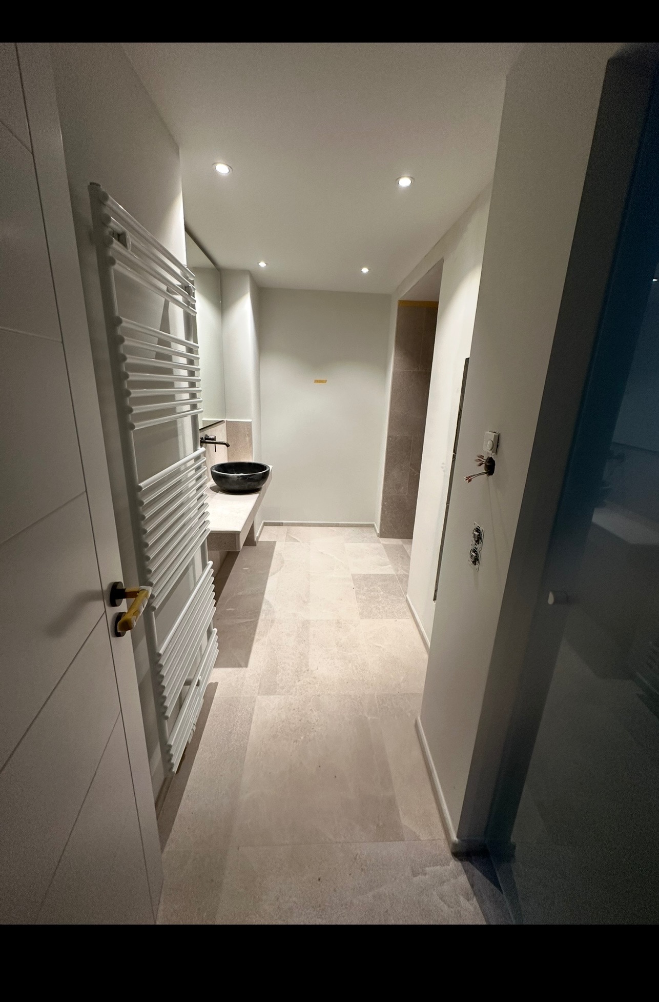 Nowoczesna łazienka z beżową podłogą z płytek, czarną umywalką i białym grzejnikiem drabinkowym. Widoczne elementy instalacji elektrycznej.