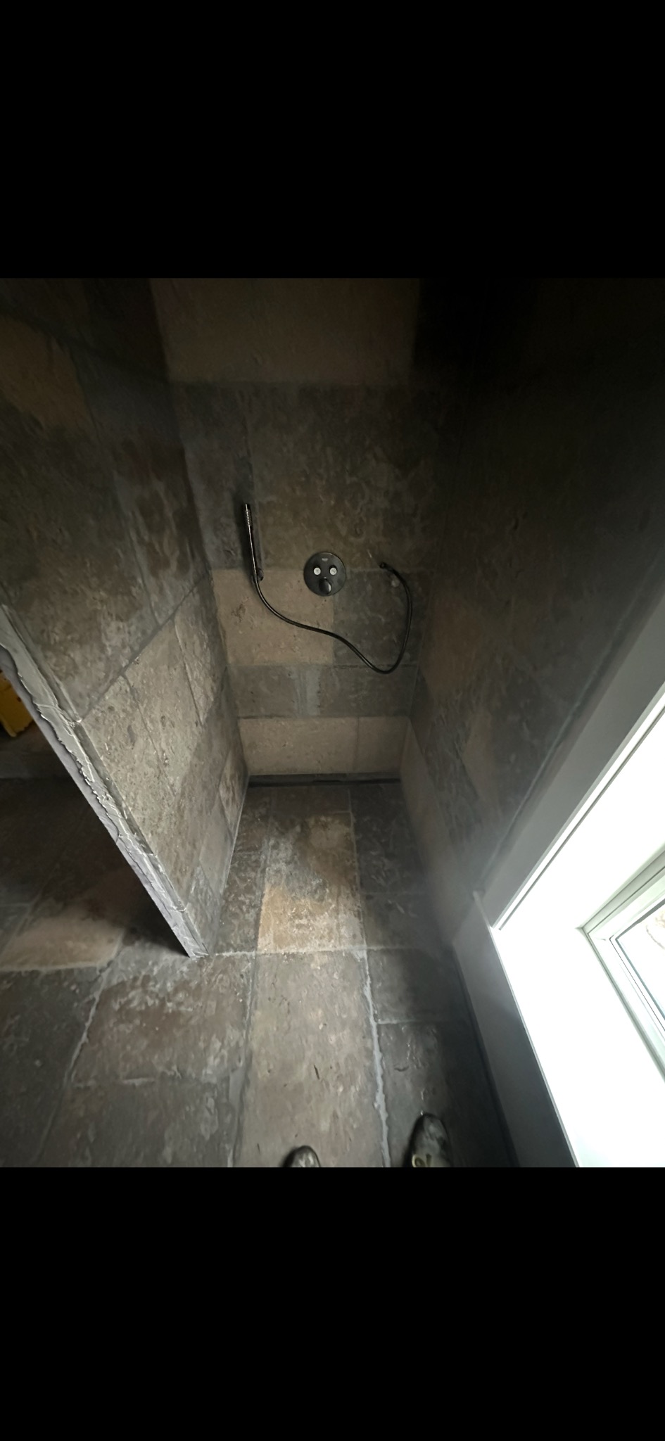 Wykończona kabina prysznicowa z szarymi płytkami imitującymi kamień, widoczny odpływ liniowy i bateria prysznicowa z wężem. U dołu kadru stopy osoby fotografującej.