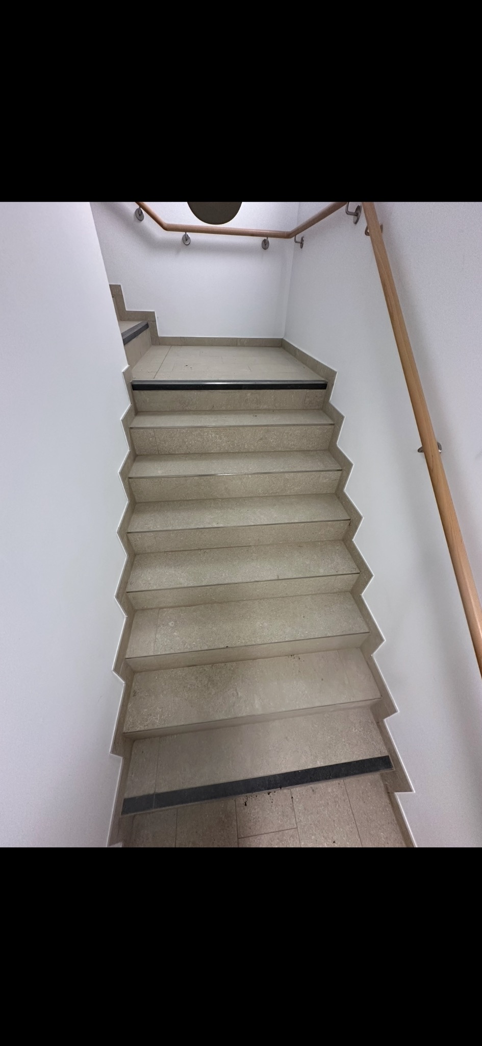 Klatka schodowa z jasnymi, wyłożonymi płytkami schodami i drewnianą poręczą. Widok z góry, ukazujący zygzakowaty kształt schodów i minimalistyczny design.