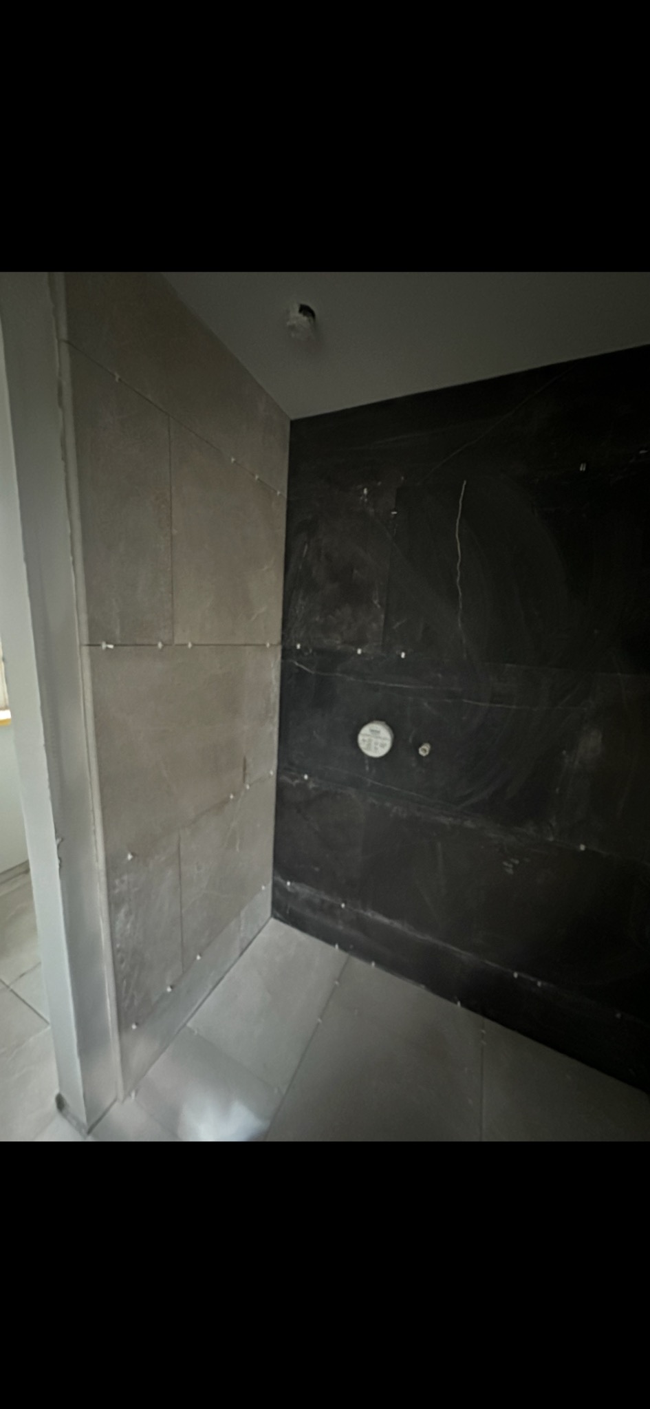 Ściana łazienki w trakcie wykańczania: szare płytki na jednej ścianie, czarne na drugiej, widoczne krzyżyki dystansowe. Ujęcie z dołu, sufit z oświetleniem.