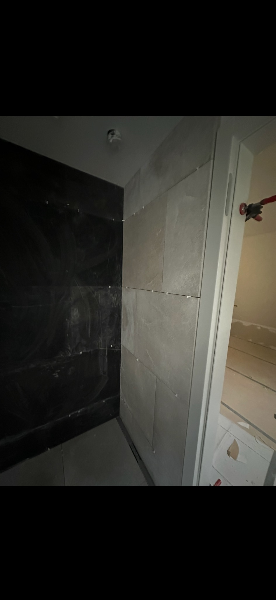 Ściana łazienki w trakcie wykańczania: część z ciemnymi płytkami, część z jasnymi, widoczne krzyżyki dystansowe i narzędzie glazurnicze przy futrynie.