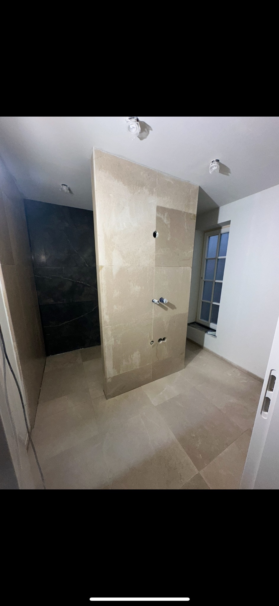 Łazienka w trakcie wykańczania: beżowe płytki na podłodze i ścianach, widoczne otwory na armaturę, ciemna strefa prysznicowa. Okno z naturalnym światłem.