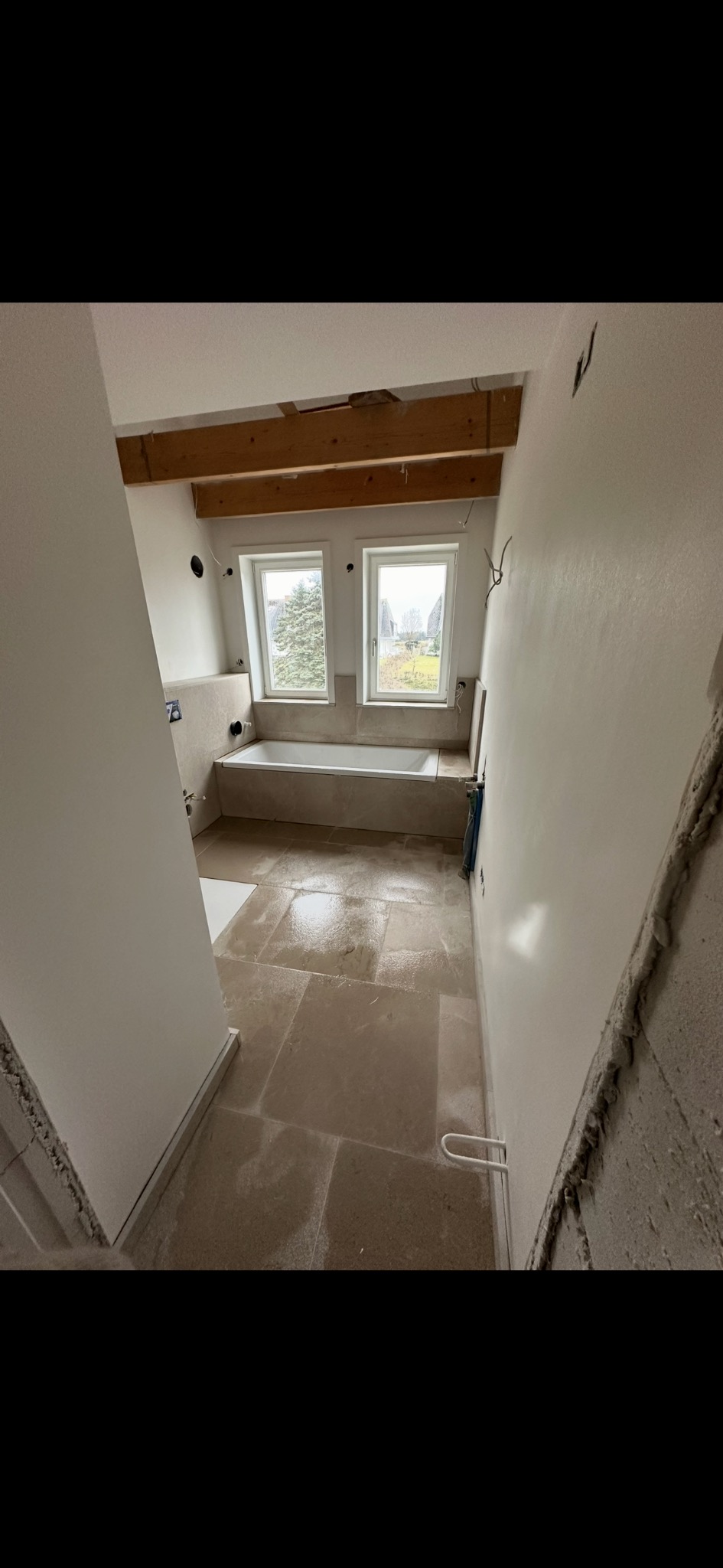 Jasna łazienka w trakcie remontu: beżowe płytki podłogowe, biała wanna pod oknami, surowe ściany. Widoczne drewniane belki stropowe i elementy instalacji.