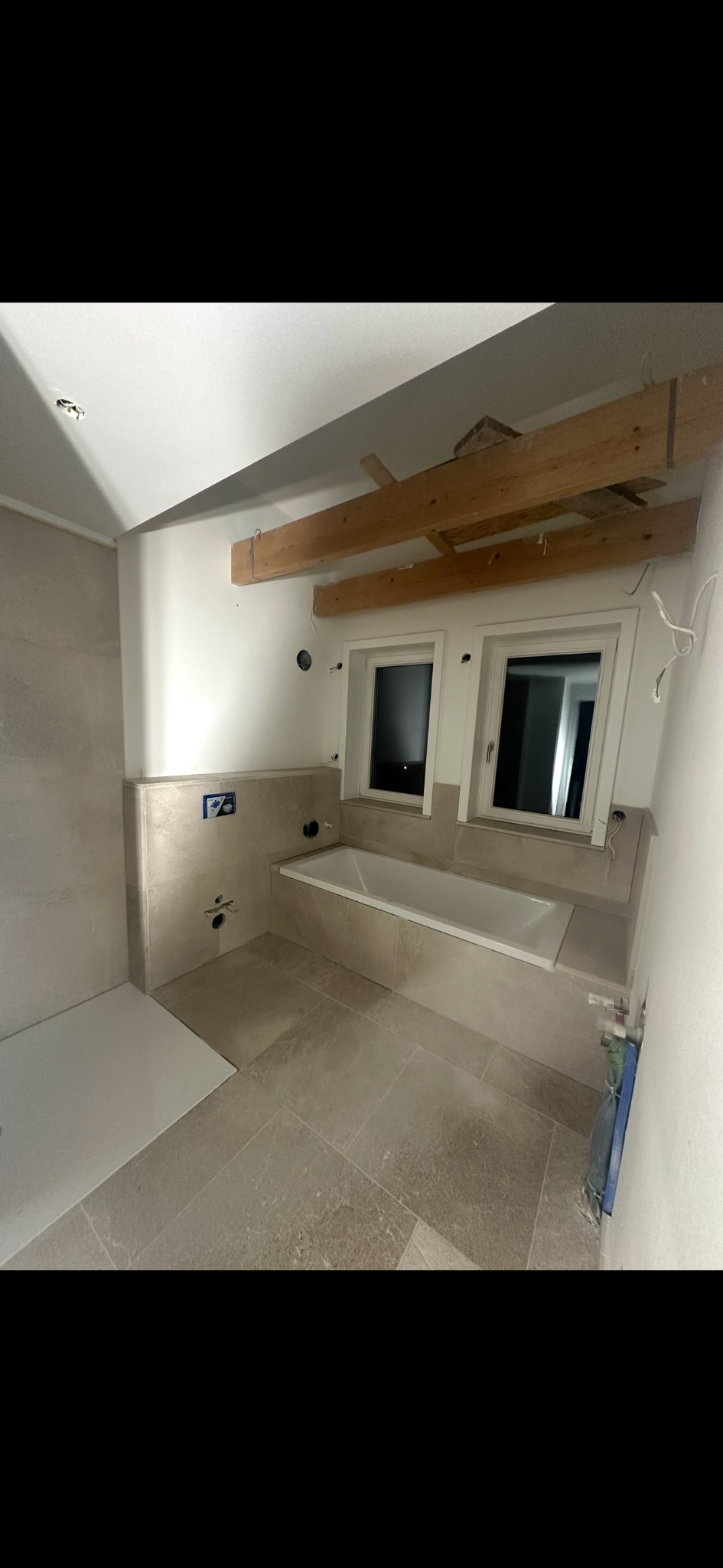 Wnętrze łazienki w trakcie wykończenia: beżowe płytki na podłodze i ścianach, wanna pod oknami, widoczne drewniane elementy konstrukcyjne pod sufitem.
