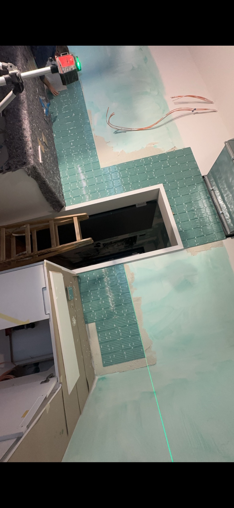 Montaż turkusowej mozaiki w łazience, widoczny laser krzyżowy, drabina i fragment szafki. Prace w toku, ściana częściowo pokryta klejem.