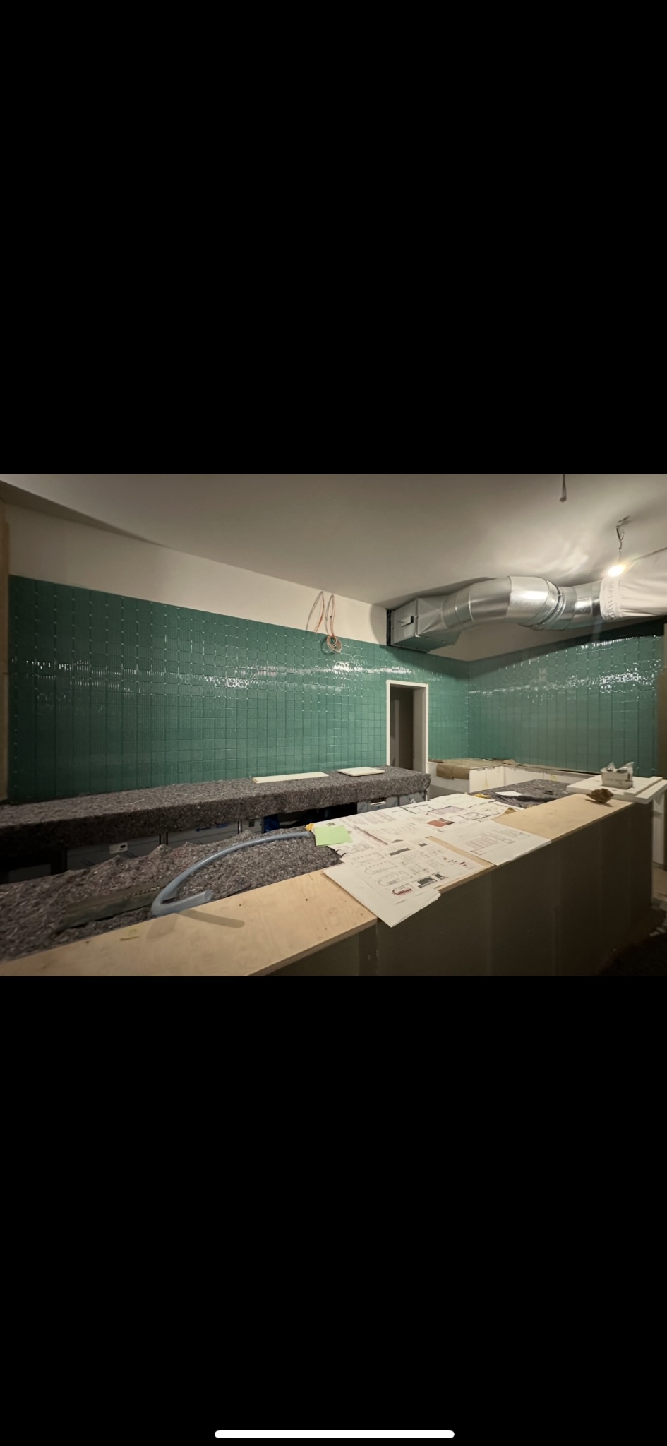 Ściany wyłożone błyszczącą, zieloną mozaiką w trakcie prac wykończeniowych. Widoczne plany i materiały budowlane na blacie. Elementy wentylacji pod sufitem.