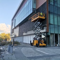 Phu - Pracownik w żółtym podnośniku koszowym, montujący elewację na nowoczesnym biurowcu z licznymi przeszkleniami, widoczne elementy konstrukcyjne i częściowo zamontowane panele.