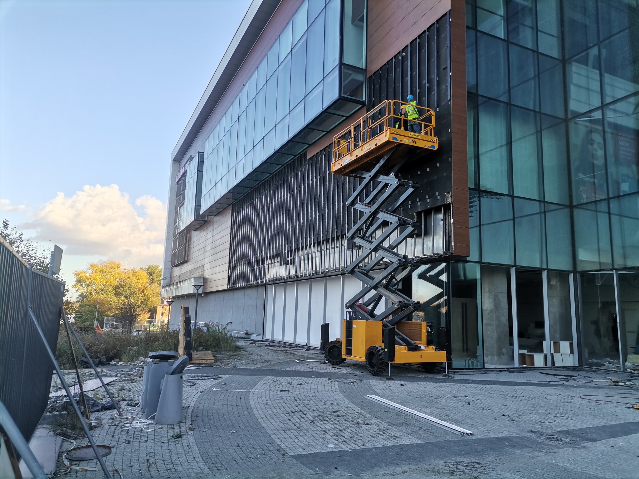 Pracownik w żółtym kombinezonie i kasku na podnośniku koszowym, montujący elementy elewacji na budynku biurowym z dużymi oknami.