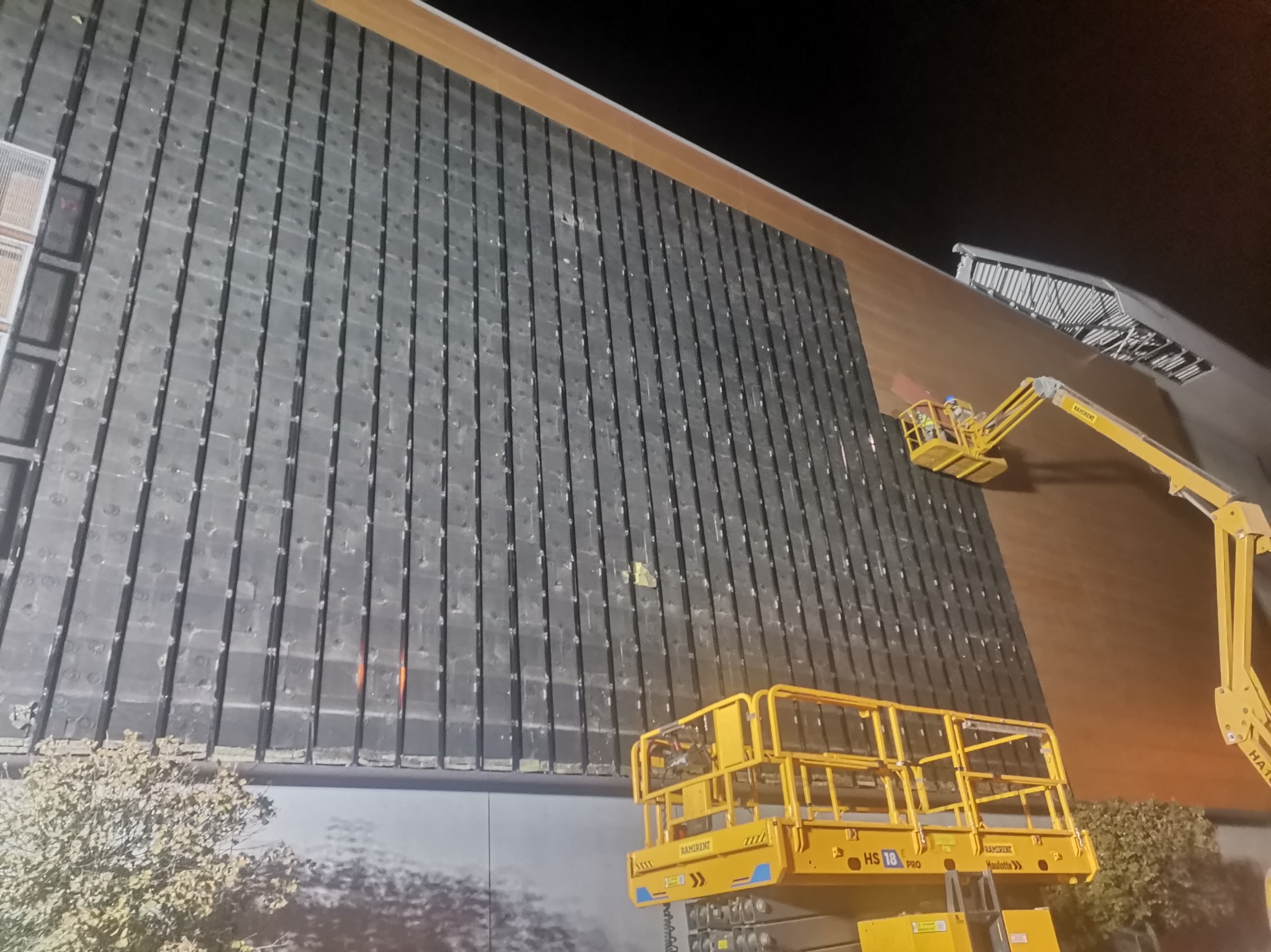 Prace na elewacji budynku z wykorzystaniem podnośnika koszowego, widoczne pionowe panele elewacyjne i pracownik w koszu podnośnika przy górnej części budynku.