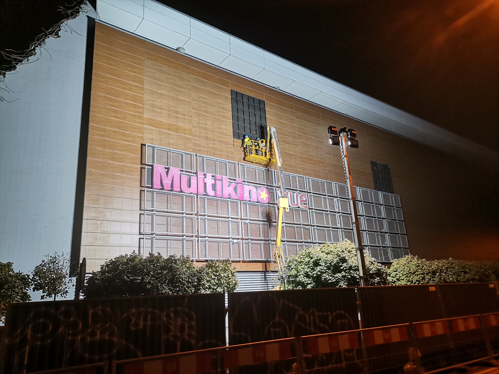 Montaż paneli elewacyjnych na budynku kina Multikino z użyciem podnośnika koszowego, widoczne ogrodzenie budowy i oświetlenie