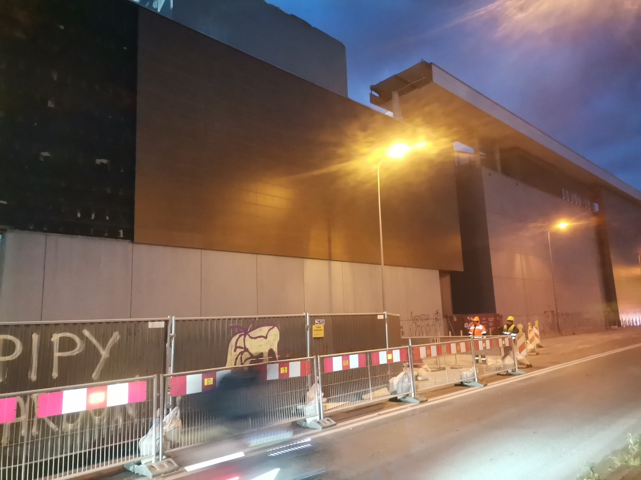 Prace budowlane przy dużym budynku, widoczne ogrodzenie z graffiti, dwóch pracowników w odblaskowych ubraniach i kaskach oraz oświetlenie uliczne.