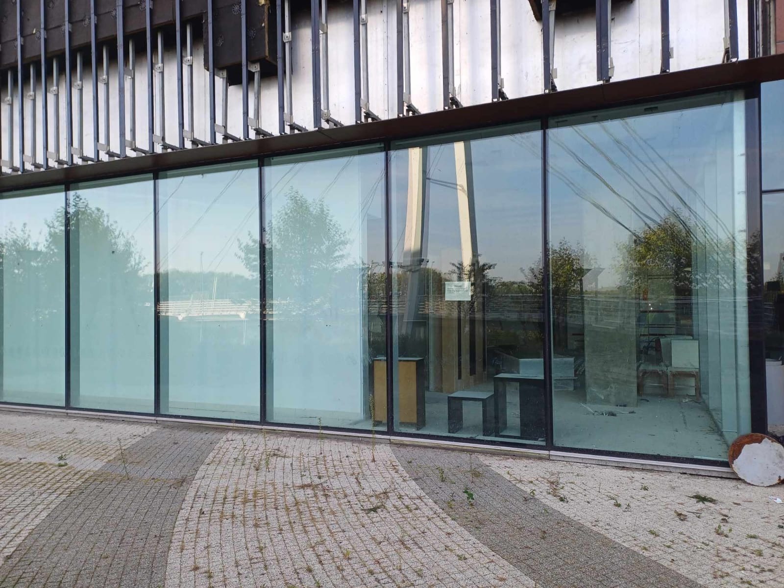 Szklana fasada budynku z widocznymi elementami konstrukcyjnymi i odbiciem otoczenia, w tym drzew i mostu; na chodniku widoczne płyty chodnikowe i fragmenty gruzu.