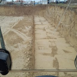 Andrzej Tynecki, usługi koparką - Głęboki wykop w piasku widziany z kabiny koparki, ślady opon na dnie, w tle zabudowa jednorodzinna i lekko pochmurne niebo.