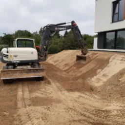 Biało-czarna koparka Terex TW110 w Jabłonnie Drugiej podczas prac ziemnych przy nowoczesnym domu z dużymi oknami, widoczne ślady opon na piasku.