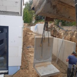 Andrzej Tynecki, usługi koparką - Montaż prefabrykowanego elementu betonowego fundamentu przy pomocy koparki na placu budowy, widoczny fragment budynku z białych bloczków.