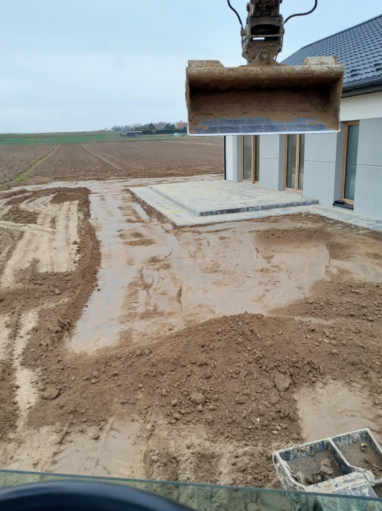 Prace ziemne w trakcie realizacji: koparka z łyżką nad mokrym, wyrównanym terenem przy domu z ułożonym tarasem z kostki brukowej, widok z kabiny operatora.