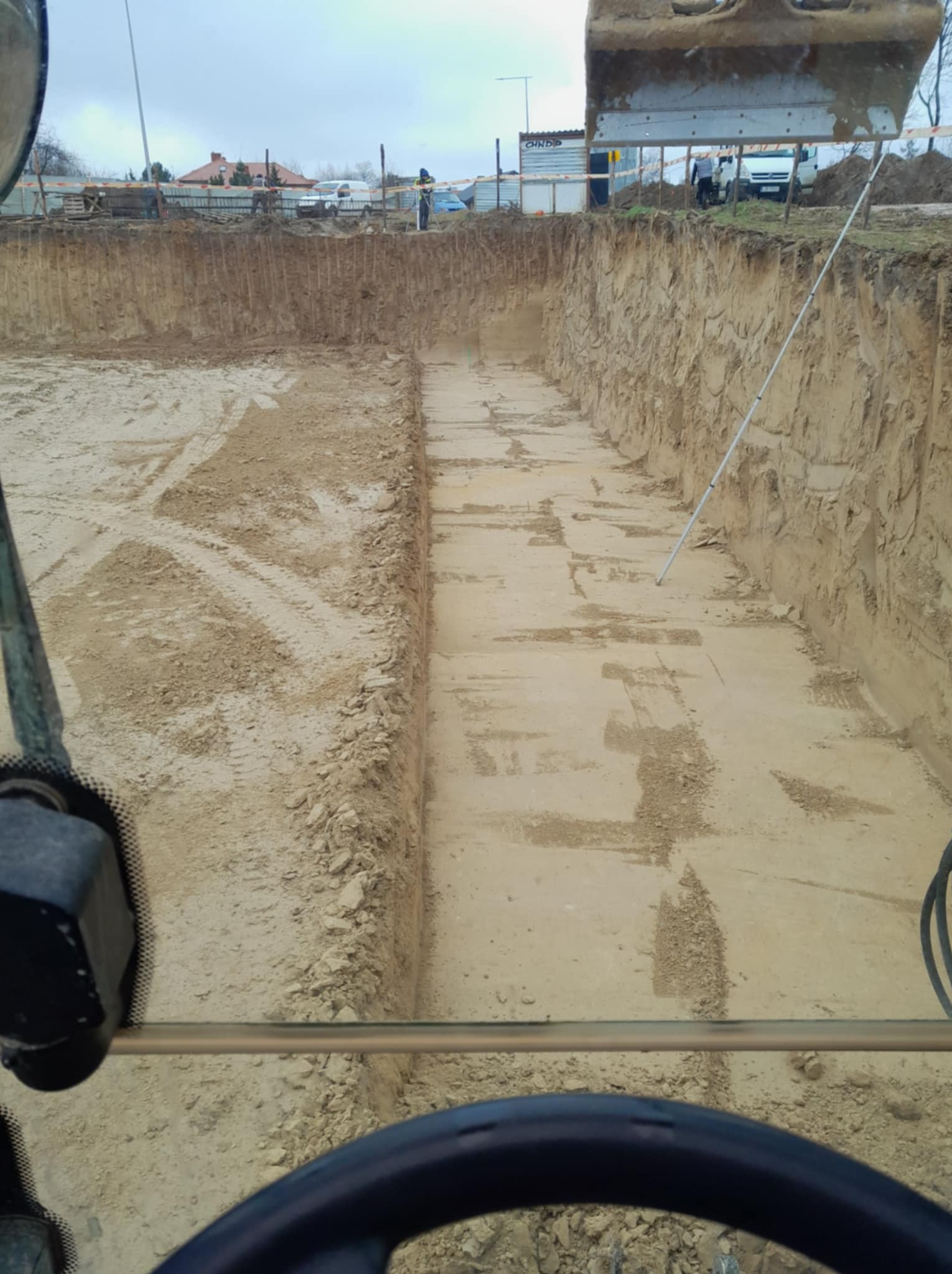 Głęboki wykop w piasku widziany z kabiny koparki, ślady opon na dnie, w tle zabudowa jednorodzinna i lekko pochmurne niebo.