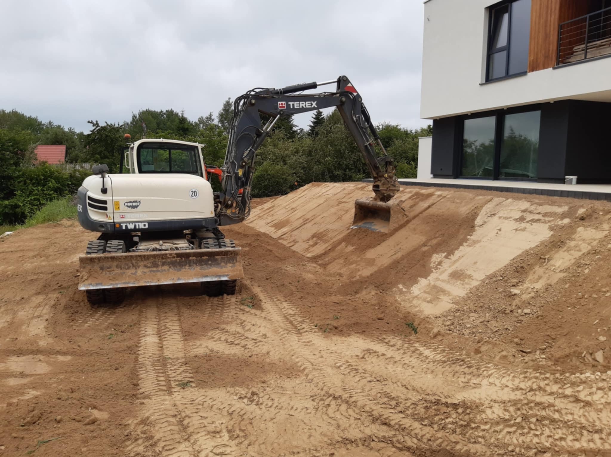 Biało-czarna koparka Terex TW110 w Jabłonnie Drugiej podczas prac ziemnych przy nowoczesnym domu z dużymi oknami, widoczne ślady opon na piasku.