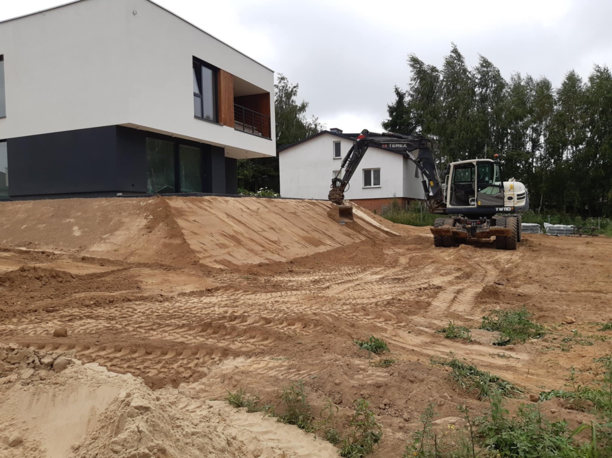 Koparka Terex na nierównym terenie piaszczystym przed nowoczesnym domem z białymi ścianami i czarnym cokołem, w tle drugi dom i drzewa.
