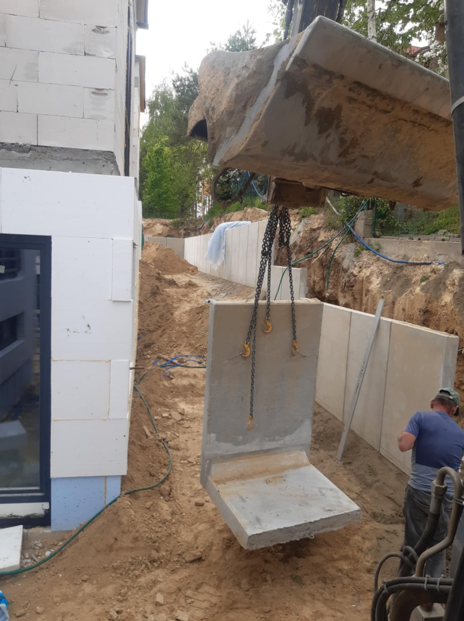 Montaż prefabrykowanego elementu betonowego fundamentu przy pomocy koparki na placu budowy, widoczny fragment budynku z białych bloczków.