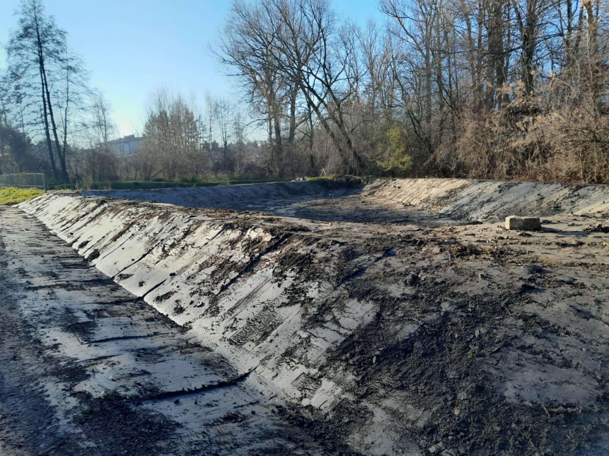 Wykonany wykop o trapezowym przekroju, zabezpieczony folią, przygotowany pod budowę zbiornika retencyjnego na tle drzew bezlistnych i błękitnego nieba.