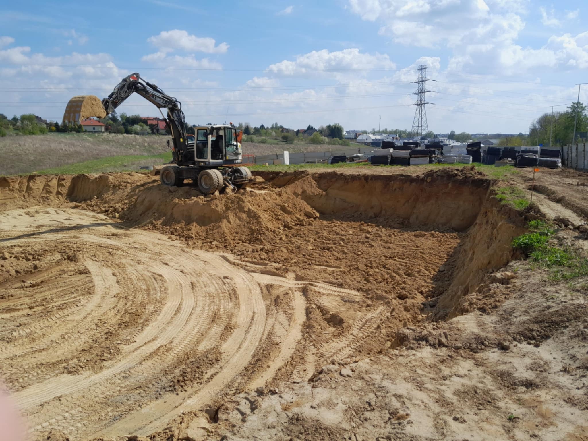 Koparka nabiera ziemię w wykopie, w tle widoczne materiały budowlane i słupy wysokiego napięcia.