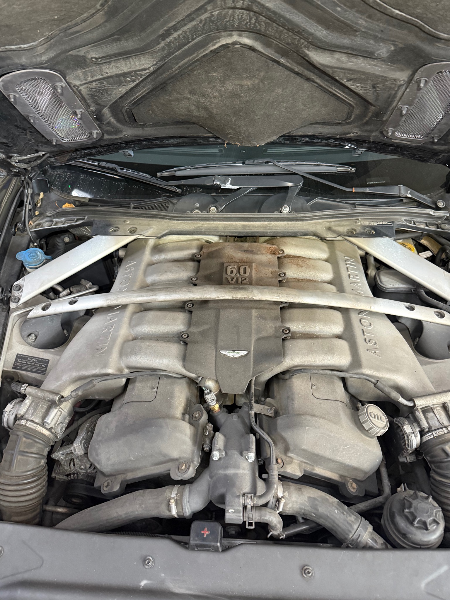 Silnik 6.0 V12 Aston Martin widziany z góry pod otwartą maską, widoczne oznaczenia modelu i logo producenta, elementy metalowe pokryte lekkim nalotem.