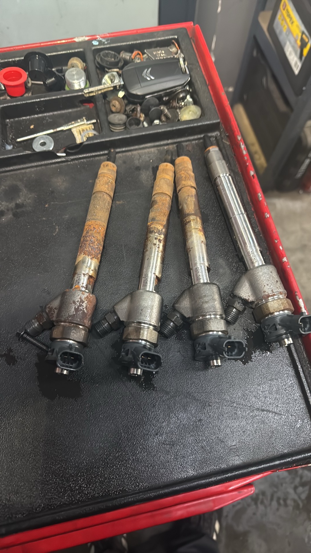 Cztery zdemontowane, używane wtryskiwacze paliwa leżące w czerwonej skrzynce narzędziowej, widoczne ślady korozji na trzech z nich, obok klucze i inne drobne części.