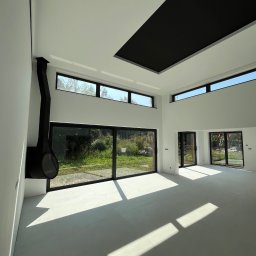 CREATE-TECH Kamil Sembol - Jasne, przestronne wnętrze z dużymi oknami i nowoczesnym kominkiem. Białe ściany i podłoga, czarny element sufitu. Widok na zieleń za oknem.