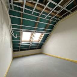 CREATE-TECH Kamil Sembol - Skośny pokój na poddaszu z oknami dachowymi. Widoczna metalowa konstrukcja sufitu podwieszanego z zieloną izolacją. Ściany pomalowane na biało, podłoga szara.