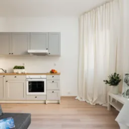 Jasna kuchnia z szarymi szafkami i drewnianym blatem, widok z salonu z szarą sofą i dekoracyjną poduszką, białe zasłony w tle.