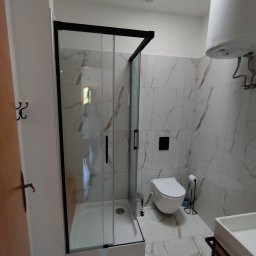 Koncept - Nowoczesna łazienka z kabiną prysznicową typu walk-in z czarnymi ramami, marmurowe płytki na ścianach i podłodze, podwieszana toaleta i biały bojler.