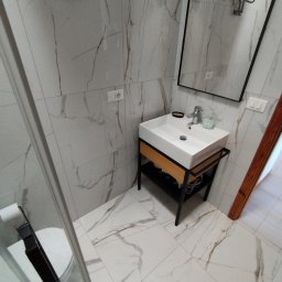 Koncept - Nowoczesna łazienka z białą umywalką na metalowym stelażu z drewnianymi elementami, lustrem w czarnej ramie i płytkami imitującymi marmur na ścianach i podłodze.