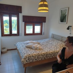 Koncept - Jasna sypialnia z łóżkiem w kwiaty, drewnianymi oknami z widokiem na zieleń i osobą siedzącą na krześle. Wnętrze dopełnia wiklinowa lampa i minimalistyczny obraz z motywem roślinnym.