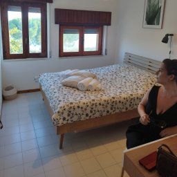 Koncept - Jasna sypialnia z drewnianymi oknami, łóżkiem w kwiaty z białymi ręcznikami i poduszkami, białą podłogą i siedzącą kobietą w czarnej sukience.