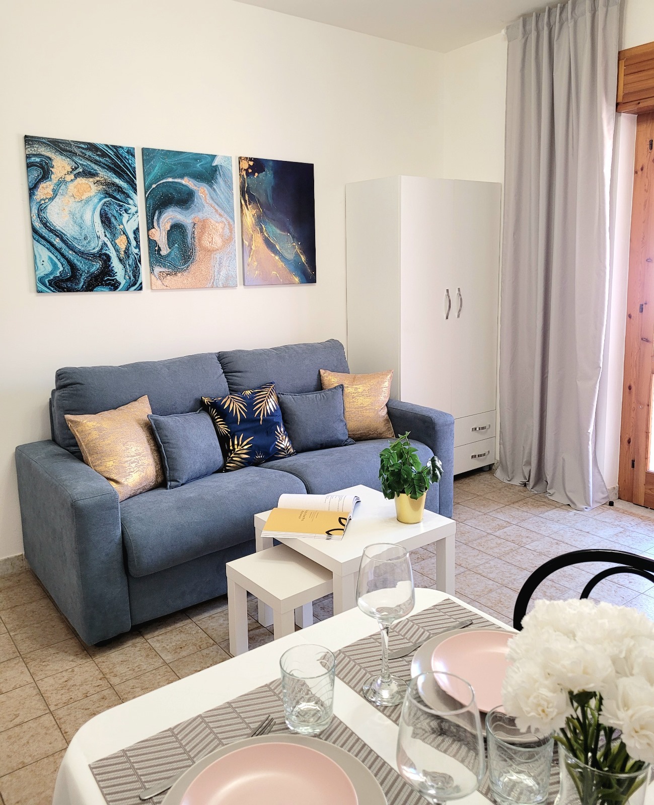 Jasne wnętrze pokoju dziennego z niebieską sofą, dekoracyjnymi poduszkami, stolikami kawowymi, białymi meblami i abstrakcyjnymi obrazami na ścianie, uzupełnione nakrytym stołem z różowymi talerzami...