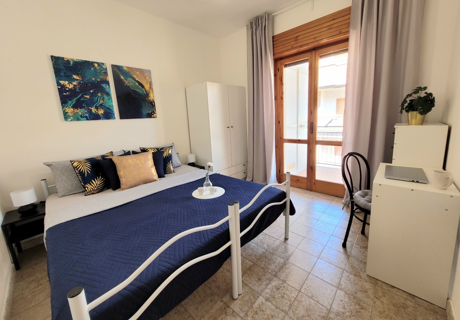 Jasna sypialnia z białym łóżkiem, granatową narzutą, dekoracyjnymi poduszkami, obrazami na ścianie, biurkiem z laptopem i balkonem z drewnianą ramą okienną.