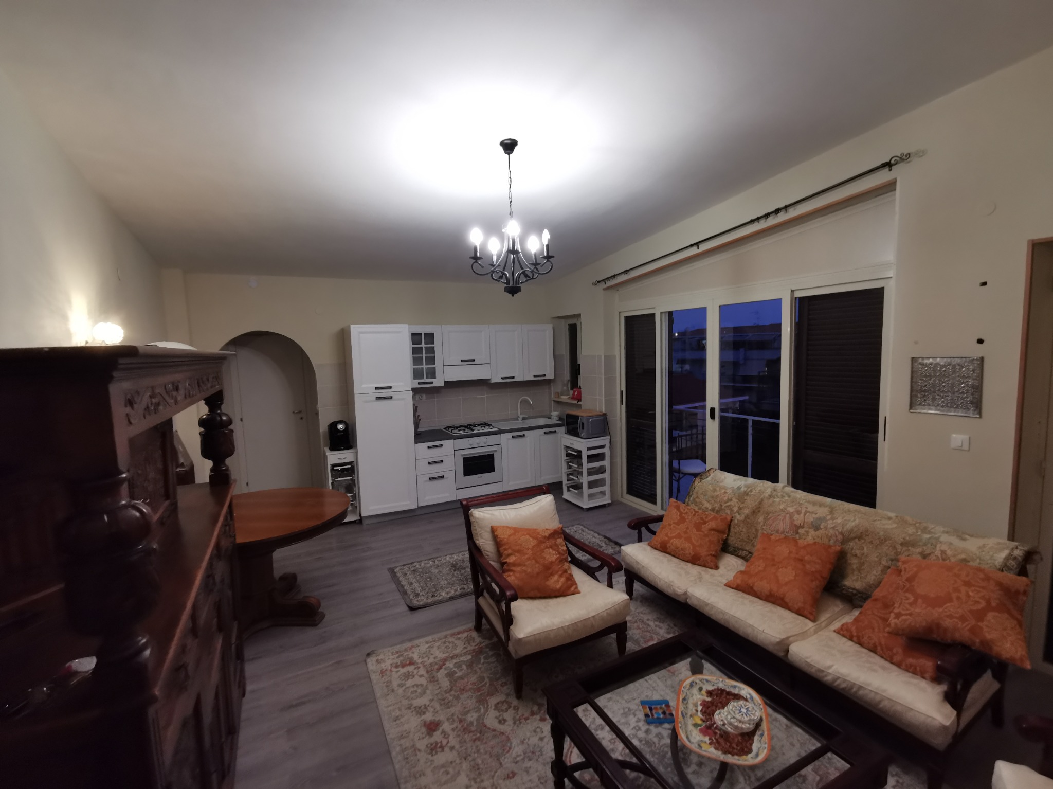 Salon z aneksem kuchennym w stylu klasycznym: białe meble kuchenne, drewniany kredens, sofa z pomarańczowymi poduszkami, żyrandol w stylu retro.