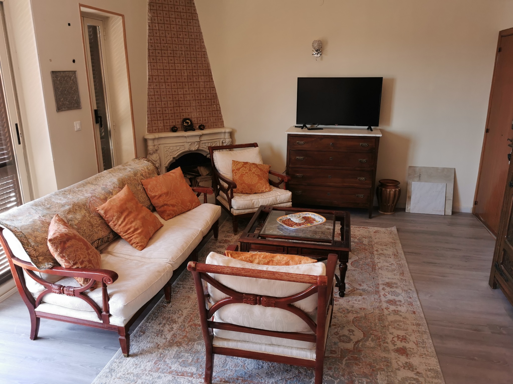 Salon w stylu klasycznym z beżowymi ścianami, drewnianymi meblami i wzorzystym dywanem; kominek obłożony kafelkami oraz telewizor na komodzie.