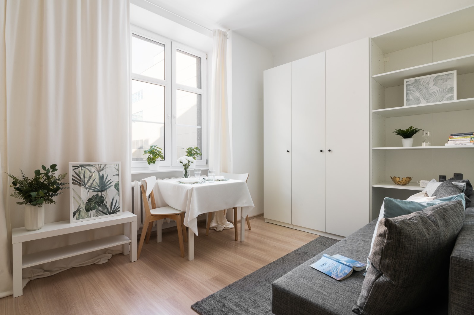 Jasne, minimalistyczne wnętrze pokoju z drewnianą podłogą, białymi meblami, stołem nakrytym białym obrusem i szarą sofą z poduszkami.
